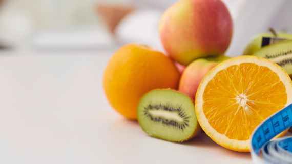 A selection of fruit on a tabletop including kiwis, oranges, apples, and a measuring tape.