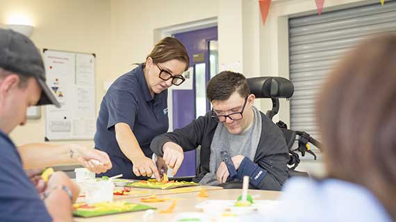 A female wearing a navy polo shirt assisting a male with learning disability with arts and crafts.