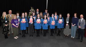 School pupils in uniform holding artwork, standing in a group along with a number of adults