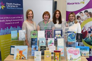 Four ladies standing in front of a table. There are books on display on the table and pop-up banners in the background