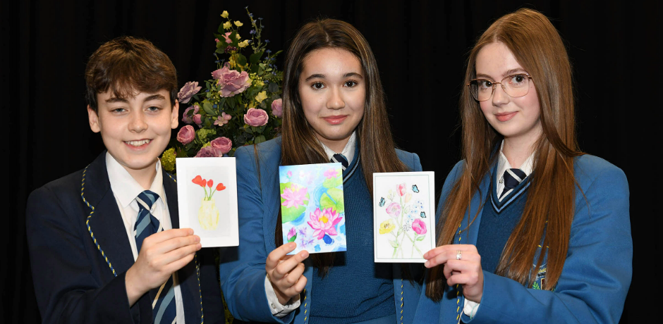 Three pupils wearing school uniform (a boy and two girls) hold up their card designs