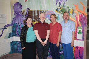 Four people standing with thumbs up with a colourful mural behind them