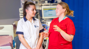 Two HSC workers standing in a ward. One is wearing a white student's tunic and the other is wearing a red tunic