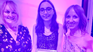 Three women in pink/purple lighting pose for a photo. One holds a certificate.