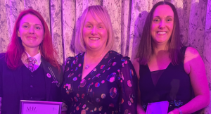 Three women in pink/purple lighting pose for a photo. One holds a trophy and another a certificate.