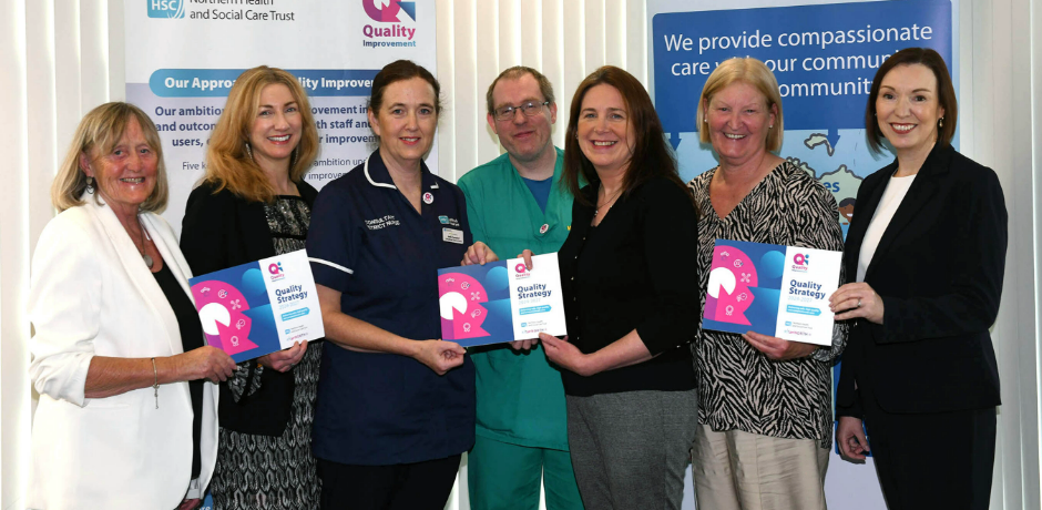 Seven people standing together with pop-up banners behind them. Some are holding a copy of the Quality Strategy document