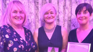 Three women in pink/purple lighting pose for a photo. One holds a trophy and another a certificate.