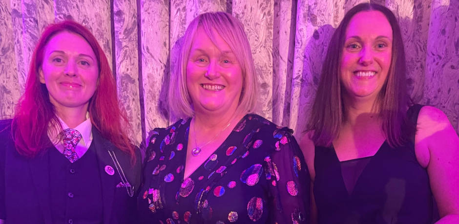 Three women in pink/purple lighting pose for a photo. One holds a trophy and another a certificate.