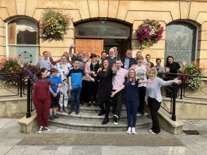 A large group of people standing together on steps outside Coleraine Town Hall, looking happy, Some are have their arms outstretched and others are waving.