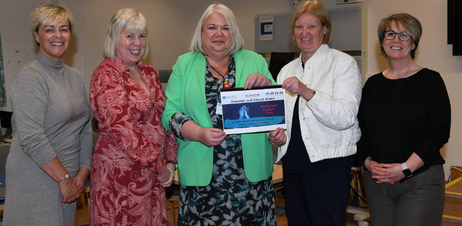 Five people standing together. Two are holding a copy of the Trust's Domestic and Sexual Abuse Workplace Support Pack.