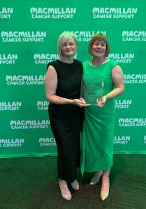 Two women standing beside each other holding a glass award. There is a green backdrop behind them which features the Macmillan Cancer Support logo.