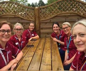 Six staff members wearing maroon uniforms sit together at a wooden outdoor table.