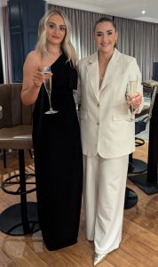 Two women standing beside each other. One is wearing a long black dress and the other is wearing a white jacket and trousers. Both are holding a glass