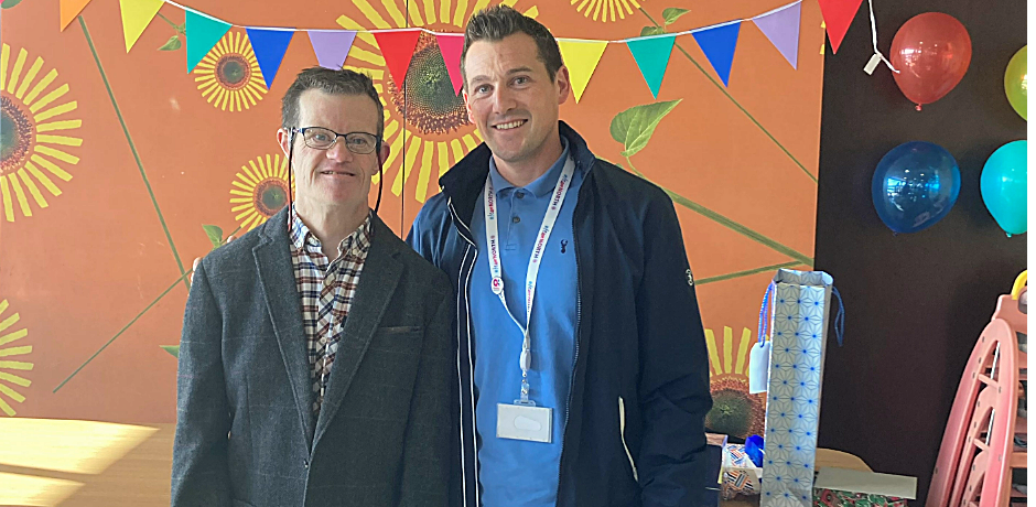 Two men standing side by side. Bunting and balloons are visible behind them.