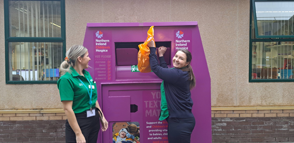 Two women standing beside an NI Hospice branded clothing bank. One is lifting an orange plastic bag into the bank.