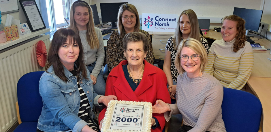 Seven people sitting together in an office. Three are in the front row, four are in the back row. There three at the front are holding a large cake which reads 'marking our 2000th referral'