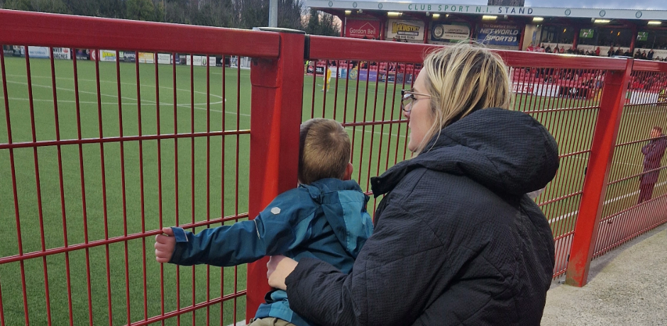 A young child looks through the fencing at the side of a football pitch, while adult sits behind him