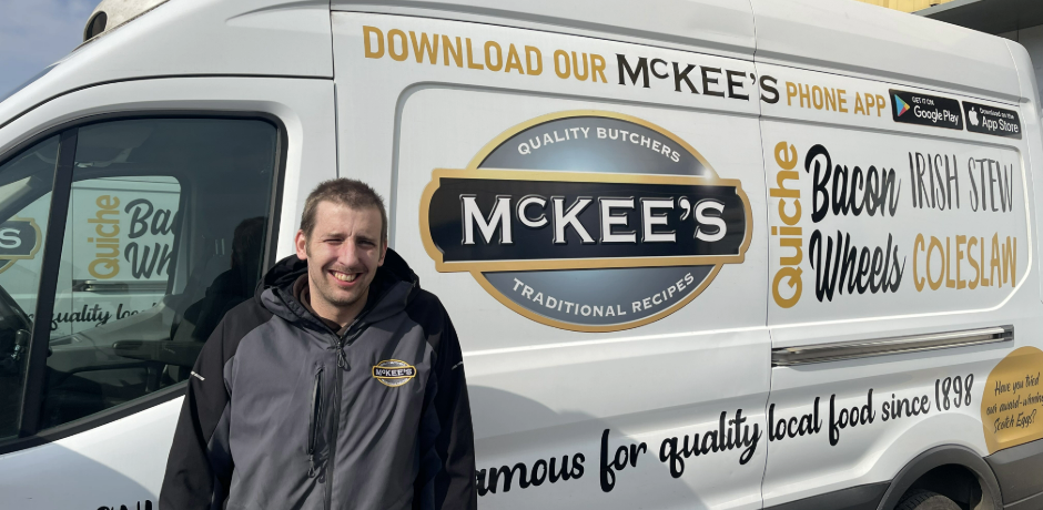 A man stands outside a white van. The man features branding for McKee's Butchers and the man is wearing a McKee's Butchers branded jacket.