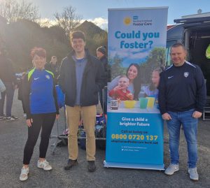 Three people stand beside a pop-up stand about fostering.