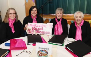 Four women sitting at a table wearing black shirts with pink scarves. They are holding a sign which reads 'celebrating rural women'.