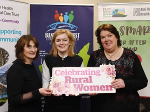 Three women standing together in front of pop-up stands, holding a sign which reads 'celebrating rural women'.