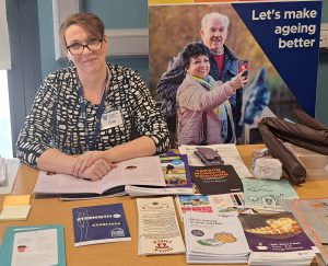 A woman sitting behind a table with pop-up stands behind. Leaflets are on the table.