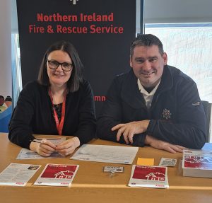 Two people sitting behind a table with pop-up stands behind. Leaflets are on the table.