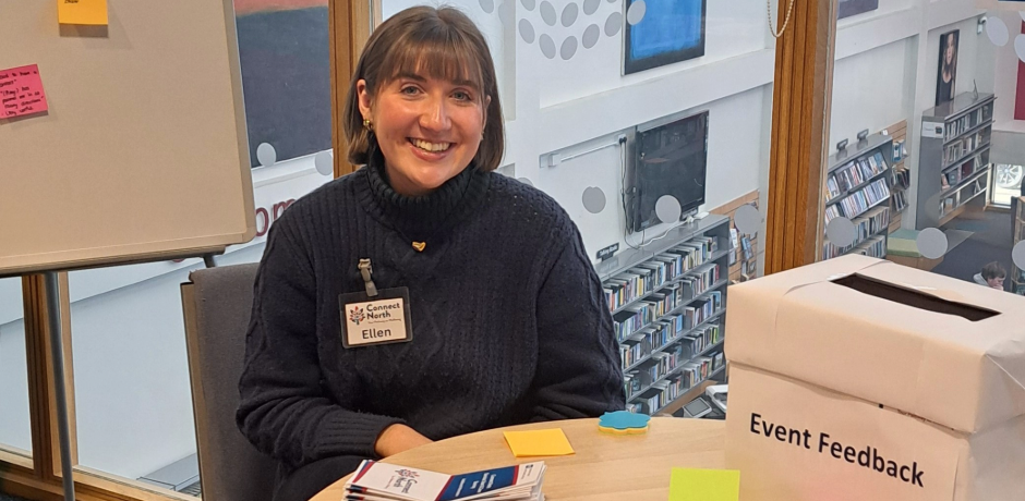 A woman sitting at a table. There is a box on the table with 'event feedback' written on the side.