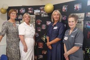 Four women standing in front of display boards. Photos and newspaper clippings are on the boards. Balloons are in the background.
