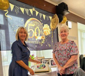 Two women stand beside a cake on a table. 50th anniversary decorations are in the background.