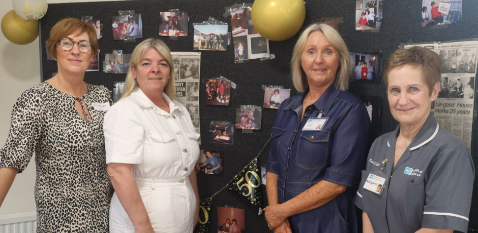 Four women standing in front of display boards. Photos and newspaper clippings are on the boards. Balloons are in the background.