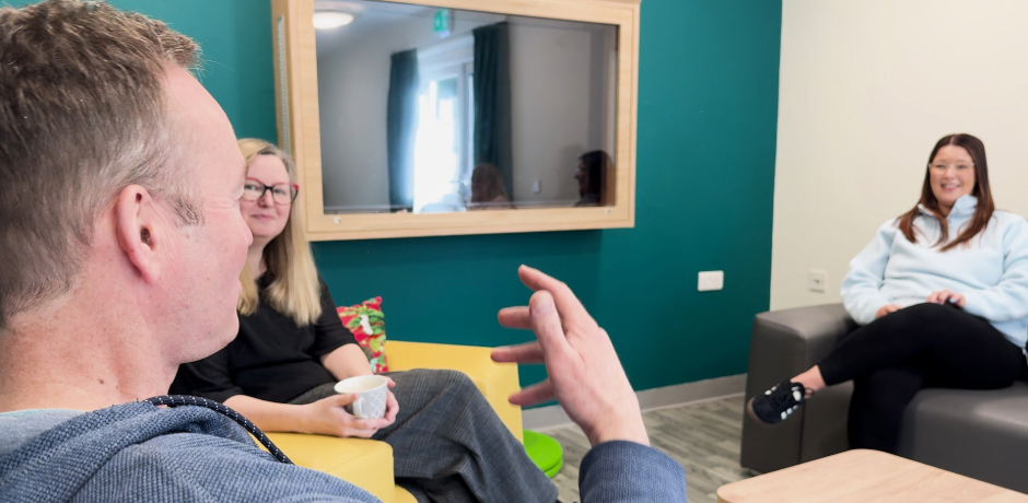 Member of a team sit on chairs and sofas chatting. Two women are in the background of the image, smiling at the the man in the foreground who is talking and emphasising with his hands.