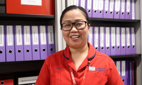A nurse in a red uniform. Behind her is a filing cabinet with a variety of coloured files lined up neatly behind her.