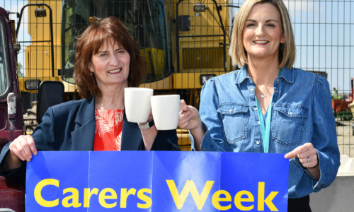 Two women stand together holding mugs and a blue banner which says Carers Week.
