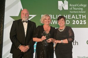 Colin Coffee wearing a tuxedo, stands beside Christina Faulkner who is wearing a black dress and holding a glass trophy. Rita Devlin is standing to the other side, wearing a black dress.