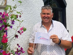 Garth Anderson standing outside holding a small white flag with Team North branding on it. A white building and purple and pink flower are in the background.