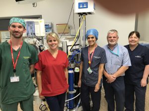 Five healthcare workers standing together. Some are wearing scrubs. Medical equipment and gas supplies can be seen in the background.