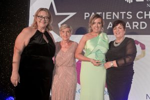 Katherine McIlroy wearing a black dress, beside Norma Sparkes who is wearing a light pink dress and Una Marie McAuley wearing a green dress and holding a glass trophy. Rita Devlin stands on the other side wearing a black dress.