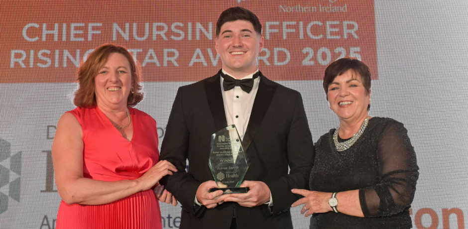 Maria McIlgorm wearing a red dress, standing with Aaron Smyth who is wearing a tuexedo and holding a glass trophy, and Rita Devlin who is standing to the other side wearing a black dress.