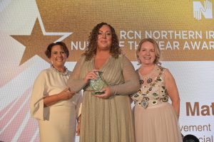 Orla Mathews stands on stage holding a glass trophy beside Nicola Ranger to the left and Briege Quinn to the right.