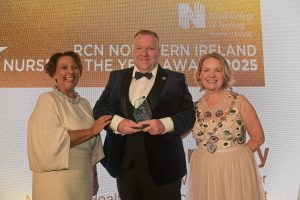 Steven Kelly, wearing a tuxedo and holding a glass trophy, stands on stage beside Nicola Ranger to the left and Briege Quinn to the right.