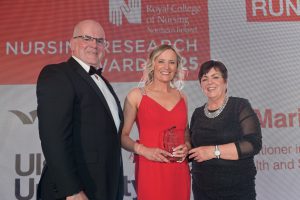 Owen Barr wearing a black tuxedo stands with Maria Betts who is wearing a red dress and holding a glass trophy, along with Rita Devlin who is wearing a black dress and stands to the right.