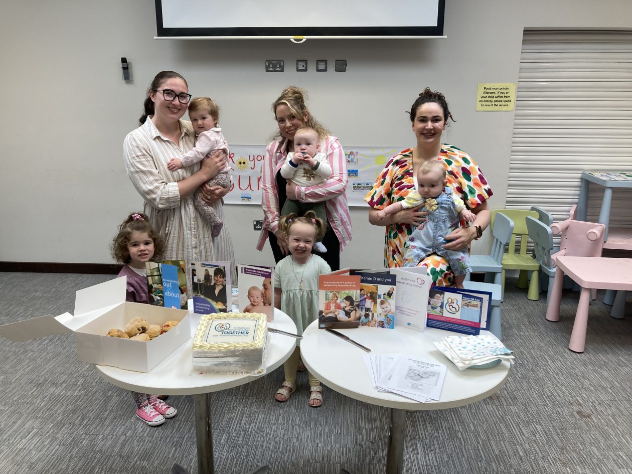 Three mums holding their babies with two toddlers standing beside them. Two tables are in front, with buns, cakes and information leaflets.