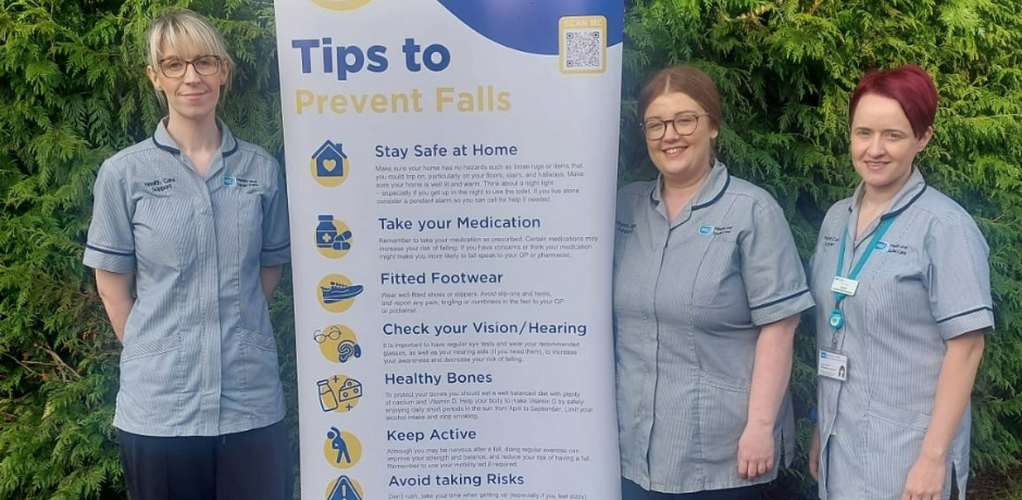 Three healthcare workers wearing grey uniform tunics, standing beside a pop-up featuring information about falls prevention.