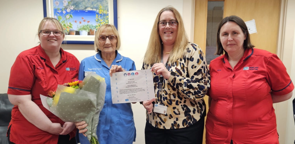 Four women stand together. Two are wearing red tunics. One is holding a bouquet of flowers and a certificate.