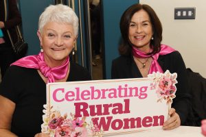 Rural Melody Choir members Lorraine Sampson and Clare Black holding a sign which reads Celebrating Rural Women. 