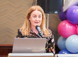 A woman speaks into a microphone, with a laptop in front of her.