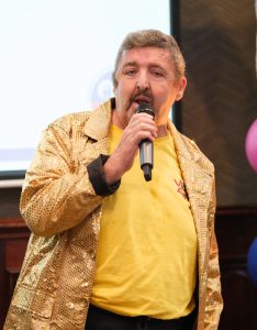 A man sings into a microphone, while wearing a gold sparkly jacket.