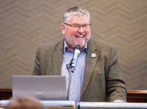 A man laughs while standing at a podium with a microphone and laptop in front of him.