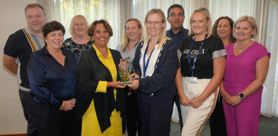 Ten people standing together. One lady in the group is receiving an award from another lady.